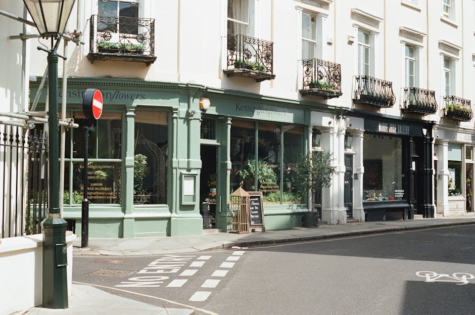 A street view of a boutique flower shop with a pastel green facade and large display windows showcasing vibrant floral arrangements. The shop, located near West Kensington station, features decorative