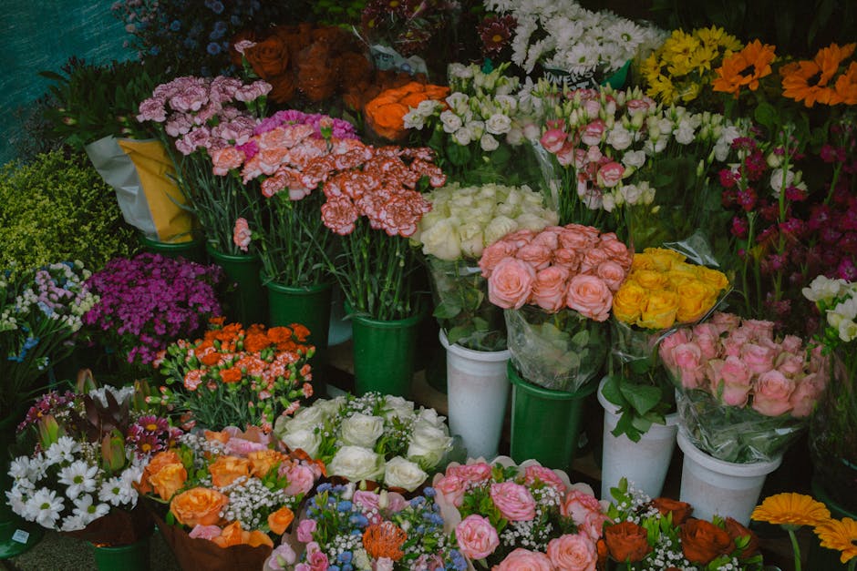 A vibrant display of fresh flowers arranged in containers and bouquets, featuring pink, white, yellow, orange, and purple blooms. The flowers include roses, carnations, daisies, and other varieties, a