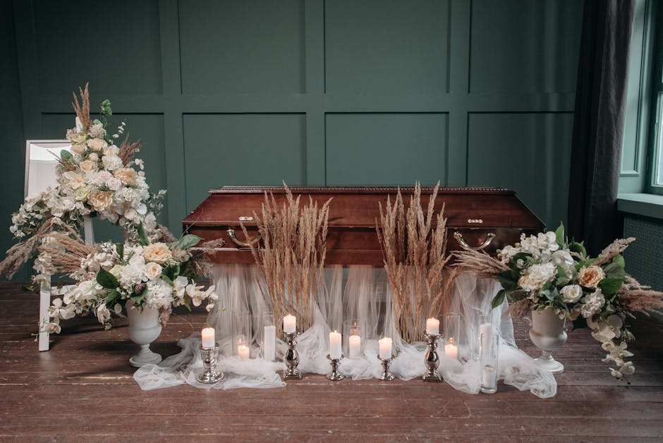A funeral floral display arranged in front of a wooden casket, featuring two large white vases filled with cream and white roses, peonies, and greenery, accented with dried pampas grass and tall wheat