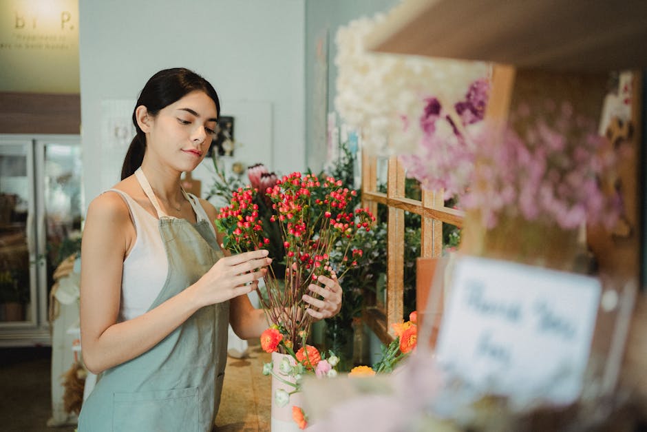 A young female florist with dark hair, wearing a beige apron over a white sleeveless top, arranging a bouquet of vibrant pink and red waxflowers in a flower shop. She stands behind a wooden counter wi