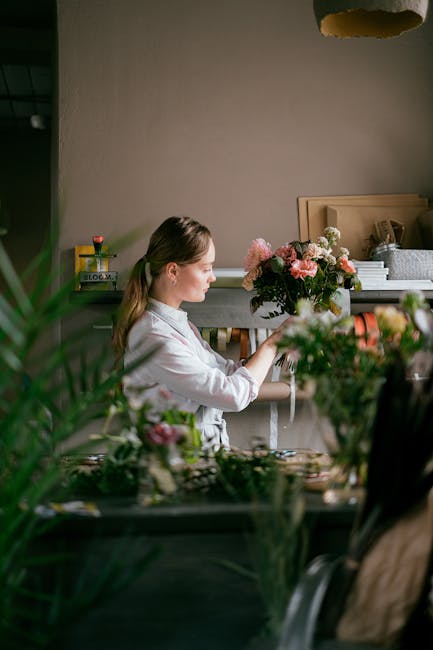 A young woman with light brown hair tied back in a ponytail arranging a floral bouquet indoors, in a cozy, well-lit room with neutral-colored walls and wooden furniture. She is wearing a light pink sh