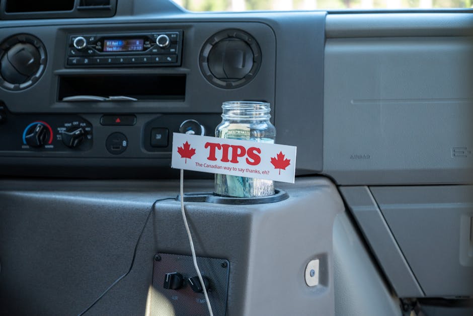 A clear glass jar filled with Canadian one-dollar coins, placed on the dashboard of a vehicle, next to a small sign with red maple leaf icons and the word 'TIPS' in bold red letters. The sign includes