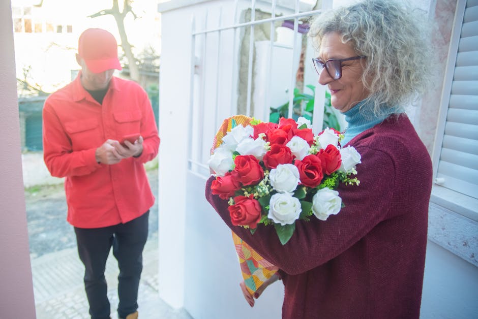 A woman with curly gray hair and glasses is holding a vibrant bouquet of fresh red and white roses, wrapped in colorful patterned paper. She is smiling as she receives the floral arrangement, which is
