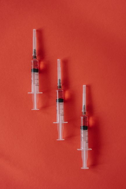 Three syringes arranged in a descending diagonal line on a plain coral-colored background, with no flowers present. The syringes are transparent with black and red markings, and are positioned upright