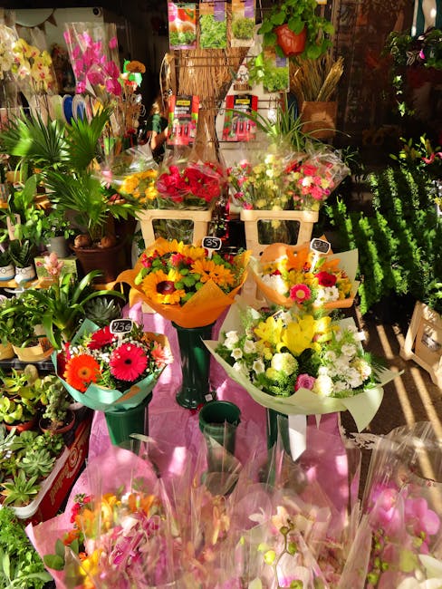 A vibrant display of fresh floral bouquets arranged on a pink cloth-covered surface inside a florist shop. The arrangement includes a mix of colourful flowers such as yellow sunflowers, pink gerbera d