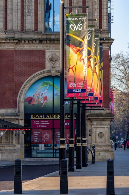 The image shows a row of black lampposts lining a paved sidewalk in front of the Royal Albert Hall, a historic red brick building with elaborate stone detailing and a large, colorful poster promoting 