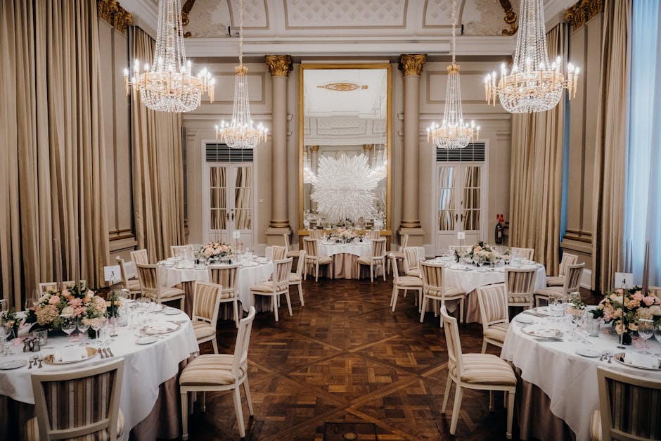An elegant wedding reception venue with high ceilings adorned with multiple large crystal chandeliers. The room features tall cream-colored drapes framing tall windows, with soft, warm lighting enhanc